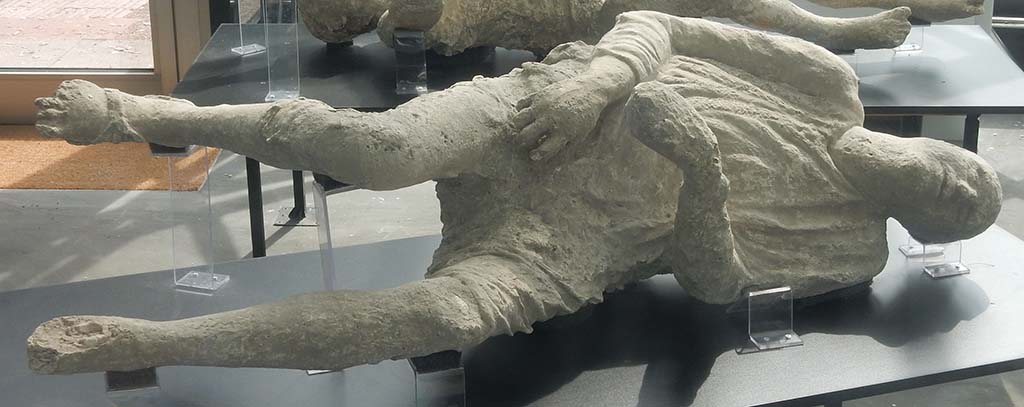 Pompeii. May 2018. General view of two more plaster-casts. Photo courtesy of Buzz Ferebee. 
In the foreground is the plaster-cast known as Victim 15, the heavily clothed man.
According to Notizie degli Scavi, (1890, P.128) the body of a man wrapped in his cloak and lying on his left side was found on 12th March 1890 in the excavations outside of the Stabian Gate. It was of a young man, of slender figure, lying on his left side, wound up in his cloak and wearing short pants which exposed his legs from above the knee. On his right foot could be clearly seen the sandal he was wearing.
See Dwyer, E., 2010. Pompeii’s Living Statues. Ann Arbor: Univ of Michigan Press. (p.107-8).