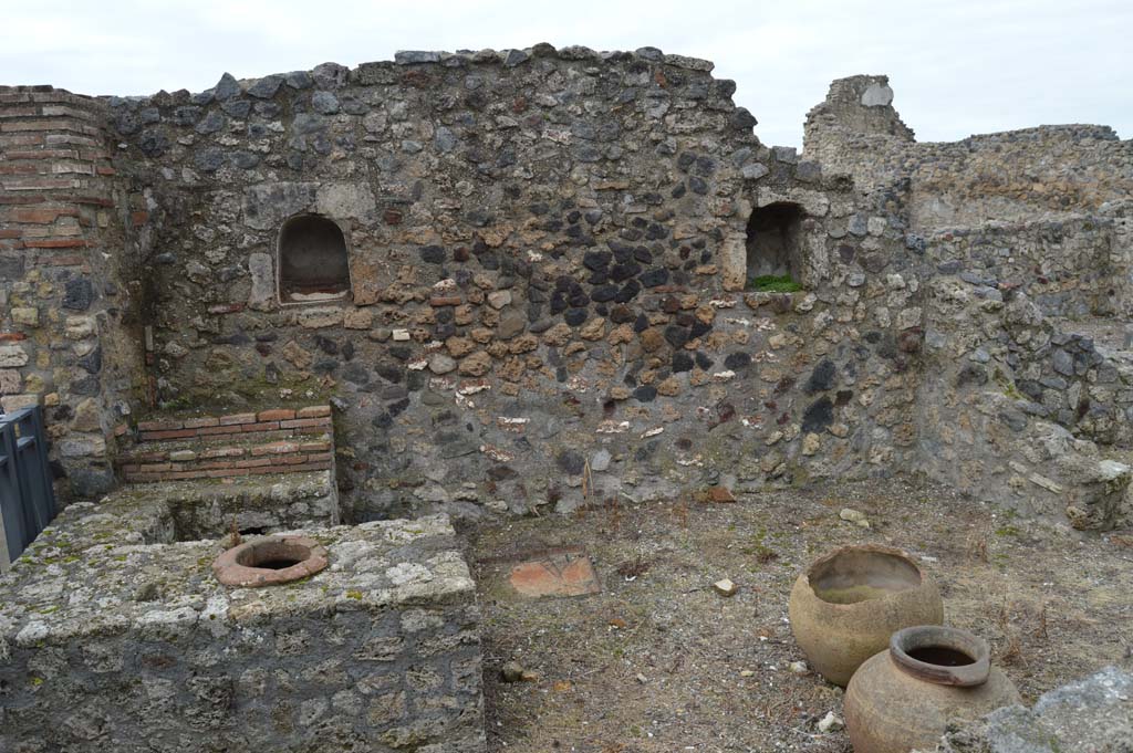 VII.16.7 Pompeii. March 2018.
Looking west towards entrance doorway, on left, counter with display shelves and inset urn/s, and west wall with two niches.
Foto Taylor Lauritsen, ERC Grant 681269 DÉCOR.
According to Boyce –
In the west wall are two arched niches, of practically the same size (h.0.46, w.0.37, d.0.28, h. above floor 1.40, distance apart 2.20), within one of which is a low step – “due nicchia per i Penati”, according to Fiorelli.
See Boyce G. K., 1937. Corpus of the Lararia of Pompeii. Rome: MAAR 14.  (p73, No.337).

