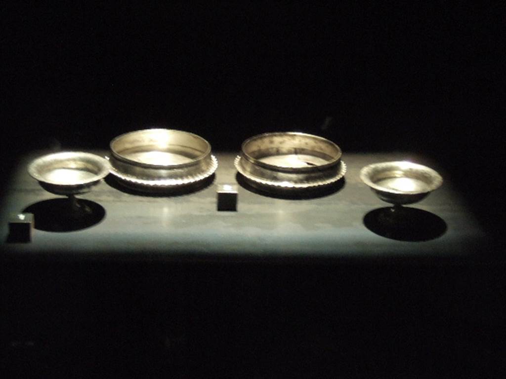 Silver found against west wall of atrium of I.6.11. 
At the edges are two goblets each in the form of a chalice.
At the centre are two bowls (ciotole) with the upper part with high edges decorated with concentric circles parallel to the rim.
Below this is a lip with an undulating ribbon pattern which separates the upper and lower parts . 
Now in Naples Archaeological Museum. 
Bowls:    inventory numbers 144804 and 144805.
Goblets: inventory numbers 144806 and 144807. 
See Guzzo, P. (A cura di), 2006. Argenti a Pompei. Milano, Electa. p.141.
