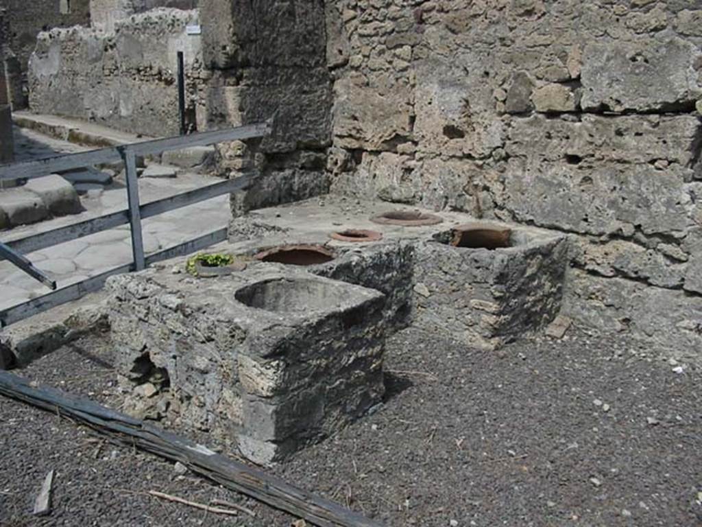 V.1.13 Pompeii. May 2003. Looking north-west at rear of counter. Photo courtesy of Nicolas Monteix.
According to Packer – 
Many small interesting finds were made in this room, including: two small bronze statuettes on bases, one of a Lar and one of Mercury, fragments of bronze candelabra, a badly preserved iron sword and two axes. The most interesting discoveries were, a human skeleton surrounded by money and jewellery: 5 pairs of gold earrings, 11 gold rings, 10 of which were set with semi-precious (?) stones, three silver rings, a small fragmentary silver shield with a head in low relief, 17 silver coins and a small silver spoon.
See Mau, in BdI, 1877, p.136.
Packer said that Della Corte had suggested that these objects represented loot collected by the man with whose skeleton they were found. Delaying his departure from Pompeii in order to plunder, he took refuge in the shop and was probably overcome by the fumes.
 See - Packer, Jim: Inns at Pompeii: a short survey. In Cronache Pompeiane, IV, 1978, (p.42-43)
