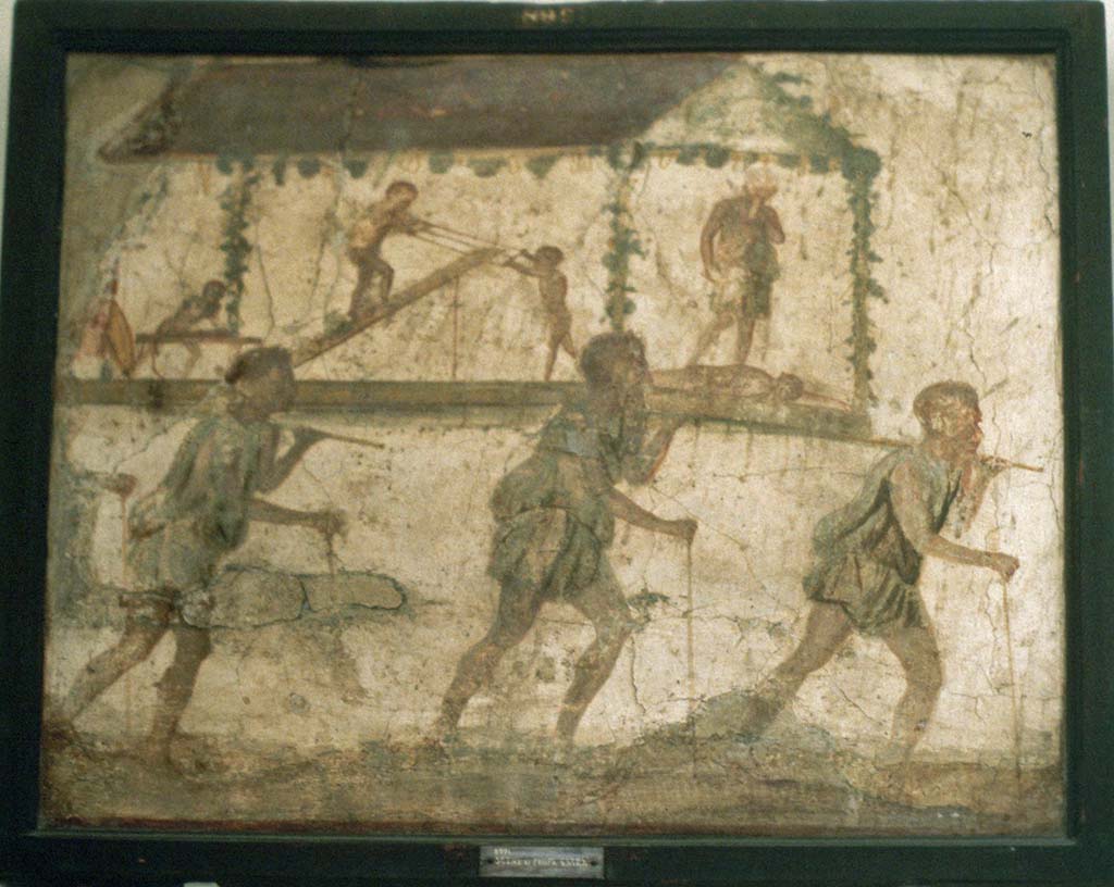 VI.7.9 Pompeii. Wall painting of the Procession of the Carpenters.   
Originally found on pilaster between entrances VI.7.8 and VI.7.9.
Now in Naples Archaeological Museum. Inventory number 8991.
See Helbig, W., 1868. Wandgemälde der vom Vesuv verschütteten Städte Campaniens. Leipzig: Breitkopf und Härtel. (1480).  
According to Leach, figures of Minerva, Mercury and Daedalus painted on its exterior pilasters (6.7.8-12), as well as another painting of a procession advertising the craft of Daedalus under the protection of Minerva and Mercury, were taken by Mau as an indication that this was a Carpenter’s workshop. 
The sign showed 3 carpenters bearing a ferculum that included a figure of Daedalus and some workmen performing carpenters’ tasks.   
A dead figure lies before Daedalus’s feet: this may well be a reference to the nephew Perdix whom the legendary artisan murdered through jealousy for his invention of the rake.   
(PPM 4, p389-91, however, suggested that the sign indicated a perfumer’s shop that dealt in spices requisite to funeral rituals). 
See Leach, E.W: The Social Life of Painting in Ancient Rome and on the Bay of Naples.
