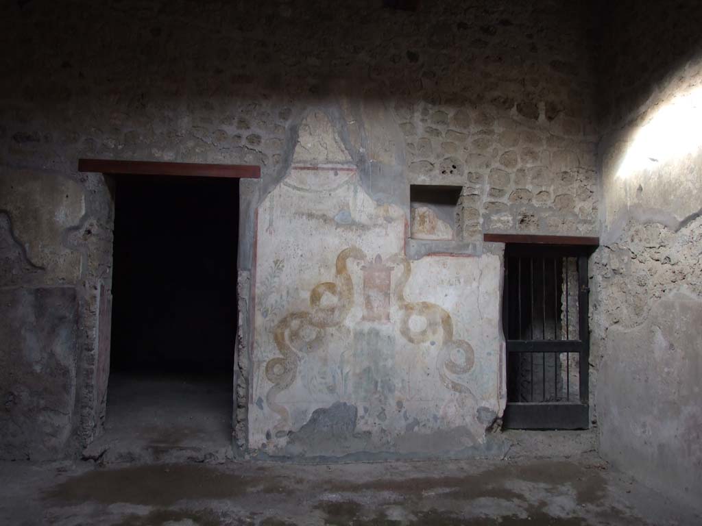 VI.16.15 Pompeii. December 2006. North wall of atrium with lararium between doorways to room I (left) and rooms K to M (right).
According to Boyce, the space between the two doorways on the north side of the atrium was painted with white stucco, bordered with red stripes.
This formed the large panel for the lararium painting.
A square niche was in the upper right-hand corner, on the rear wall of which was painted the figure of the Genius.
Hanging down on each side of him was a garland, on the side walls were painted red flowers with green leaves.
Of the two Lares originally painted on the white background of the wall, one on each side of the niche, only the lower part of the one on the left remained.
Below the figure of the Lar was painted a tall cylindrical altar, furnished with two eggs, a pine cone and fruit.
On each side was a huge yellow serpent, each coiled serpent about two metres in length.
The one on the left had a red crest and beard. The one on the right had neither.
In the background were rather large plants bearing fruit.
Over the surface of this picture had been spread a thin wash.
It would appear that the lararium belonged to an earlier decoration of the house and was to be replaced by a new one to be painted on the thin wash.
See Notizie degli Scavi di Antichità, 1908, p. 67.
See Boyce G. K., 1937. Corpus of the Lararia of Pompeii. Rome: MAAR 14. (p.58, no.224, Pl.16,1) 
See Fröhlich, T., 1991. Lararien und Fassadenbilder in den Vesuvstädten. Mainz: von Zabern. (L75, picture. 39,2)
See Giacobello, F., 2008. Larari Pompeiani: Iconografia e culto dei Lari in ambito domestico. Milano: LED Edizioni. (p.185)
