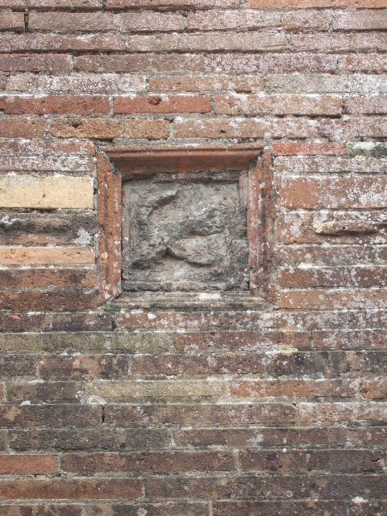 VII.2.32 Pompeii. May 2006. Plaque showing a flying phallus, on wall between VII.2.31 and VII.2.32. According to Della Corte, a characteristic, complex relief (embedded in the wall to the left of the first entrance) destined to protect the hostelry.  

