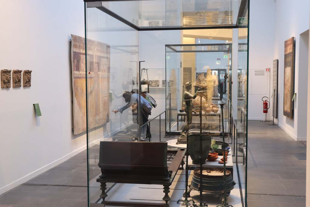 VIII.1.4 Pompeii Antiquarium. February 2021. Looking through same room across display case from in front of the statue of Nike.
Photo courtesy of Fabien Bièvre-Perrin (CC BY-NC-SA).
On the left wall are the terracotta plaques from Pompeii.
On the right wall is the second wall painting from the north wall of the triclinium of the House of the Golden Bracelet.
In the far centre, on a shelf, are the glass panels from the House of Fabius Rufus.
