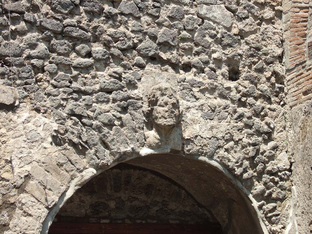 VIII.7.20 Pompeii. May 2006. West side. Head (keystone) over entrance to room under the seating.
Kuivalainen describes –
A single naked bust depicts frontally a young man with strong features. Tight curls frame both sides of the face and clusters of berries of ivy adorn the hair. Covered with stucco. (The head is damaged, stucco partly fallen, eg, in the nose and curls).
Kuivalainen comments –
Eschebach changed his mind on the identification, from a satyr to Bacchus, and with reason. The audience entering the cavea from the courtyard through the west entrance saw the bust of Bacchus above them, and were thus elevated to the right mood for the performance. The Greek features of the architecture also support the identification as Bacchus, the protector of the theatre.
See Kuivalainen, I., 2021. The Portrayal of Pompeian Bacchus. Commentationes Humanarum Litterarum 140. Helsinki: Finnish Society of Sciences and Letters, (H5, p.210).
