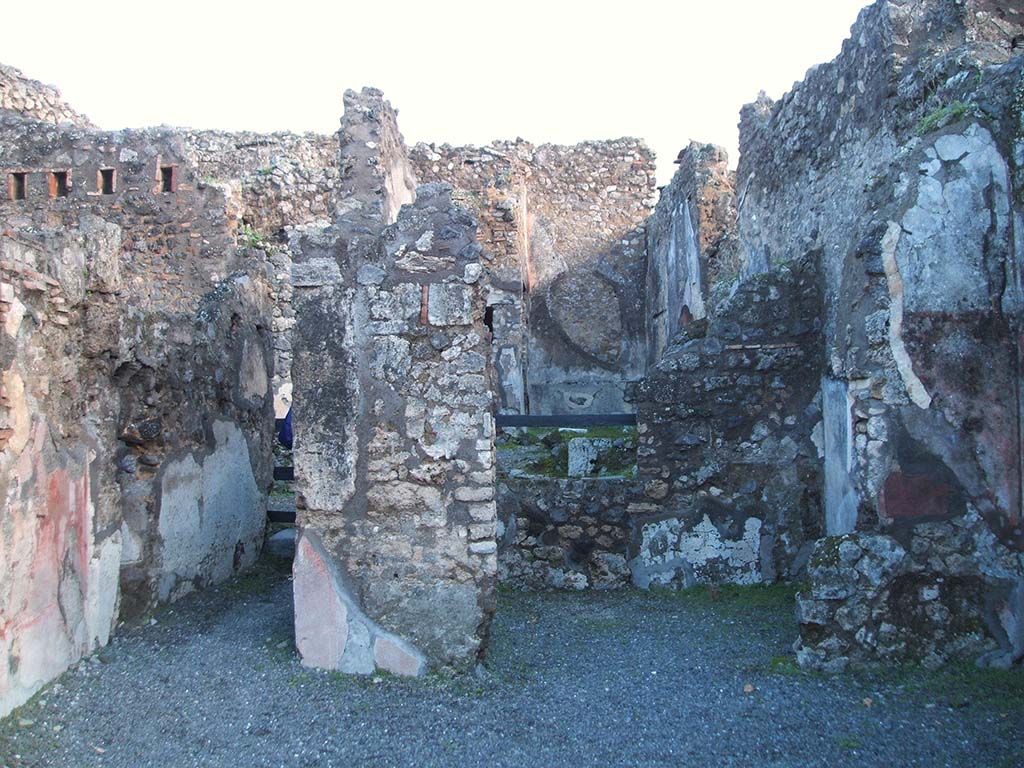 IX.2.7 Pompeii. December 2004. Looking east to rear room of shop (b) with corridor to rear (d) on north side of it.
According to Minervini –
From the shop-room, by a threshold of white marble, one entered into another small room. 
The flooring was of opus signinum with a rough mosaic of white stones in the centre.
The walls were white, with panels of different colours, decorated with masks, birds and animals: then a drum suspended by ribbons, a small half-open basket, and another uncertain object. From here and there, on the two side walls, you could see a bearded figure, with wide tunic, who held a vase in the shape of a kantharos on his head with both hands. In the wall on the right are shown two Cupids, partly faded, and as many on the wall to the left, one with patera the other with lyre, and all with fluttering cloak: many times Cupid with lyre appeared in pompeian paintings.  
At the top of the wall, there are residues of stucco composed of a flat fascia with pretty foliage, and a curve with palm leaves and stalks (caulicles): in the space comprised by these two lines a hippocampus and two dolphins are painted.
See Bullettino Archeologico Napoletano Nuova Serie: Anno Primo, no.4, Agosto 1852, p.25. Minervini.
