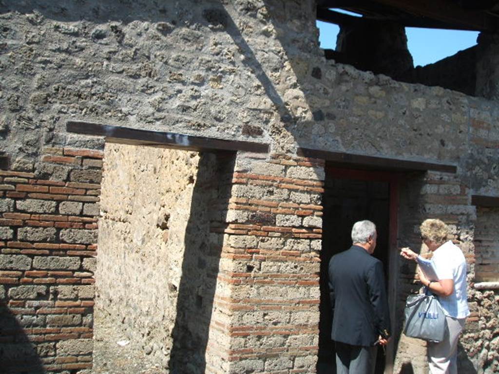 IX.5.16 Pompeii. May 2005. West side of atrium a’, on the left, doorway to kitchen and latrine e’.
On the right, doorway to cubiculum f’.
According to Boyce, on the south wall of the kitchen was a large rectangular panel of white stucco.
On it were painted two yellow serpents with red crest and beards. 
These were on either side of a tile embedded and projecting from the wall.
A fire was painted on the wall above the tile.
The background was painted with plants, and across the top were two painted garlands.
On each of the garlands was a painted bird.
See Boyce, G. K., 1937. Corpus of the Lararia of Pompeii. Rome: MAAR 14.  (p. 86, no. 425)

In IX.5.16, the first place on the left of the atrium was searched again, and found were several iron nails, some fish bones, and in terracotta: two containers for oil, a vase in fragments, two amphorae, two pot lids, and a broken bowl containing ash.
See Notizie degli Scavi di Antichità, 1885, (pp. 162-4).

