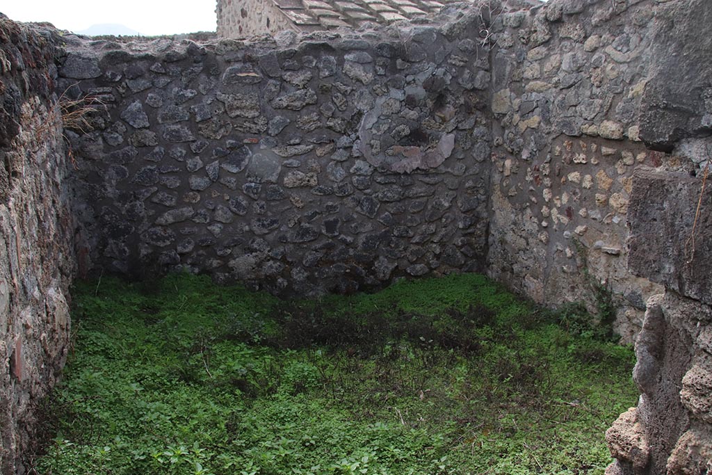 IX.7.19 Pompeii. October 2024. Room k, looking south through doorway in corridor f. Photo courtesy of Klaus Heese.
The east wall and south-east corner are on the left, presumably the site of the lararium painting, but nothing now remains.

According to Boyce, a small room, room k, located between kitchen and garden on the south side of the house, was originally part of the garden.
This small room had a doorway into the kitchen. 
Then the room was separated from the garden and the doorway to the kitchen was bricked-up.
On the east wall of this new room was a lararium, with a Genius standing to the right of a tripod.
On the other side of the tripod stood the tibicen. Below and to the right, ran a camillus. 
Below this were two serpents, gliding, one from each side, amongst plants towards an altar.
Between this painting and the south-east corner of the room were painted, two pots, a large bottle, sausages, a calf’s head, ribs of pork on a spit, and a phallus.
He references – Not. Scavi, 1880, 491; Bull. Inst., 1883, 82.
See Boyce G. K., 1937. Corpus of the Lararia of Pompeii. Rome: MAAR 14. (p.88, no.438) 
According to NdS, the were also two Lares:
To right in the atrium there is a room communicating with the back room of the following taberna, and the access to the kitchen with the latrine, and to a cell, where the lararium painting is divided into two strips; in the lower part there are the two serpents, which rush towards the altar with the offerings, and in the upper part the sacrificing Genius Familiaris, with tibicen and a camillus between the two Lares.
See Notizie degli Scavi di Antichità, 1880, p. 491.
Fröhlich notes: Boyce does not mention Lares.
Boyce 438 (IX 7, 19) describes the Genius with Camillus and Tibicen grouped around the altar in the usual way, but without the Lares. 
See Fröhlich, T., 1991. Lararien und Fassadenbilder in den Vesuvstädten. Mainz: von Zabern. (p.112).
See Giacobello, F., 2008. Larari Pompeiani: Iconografia e culto dei Lari in ambito domestico. Milano: LED Edizioni, (p.209, no.106)
