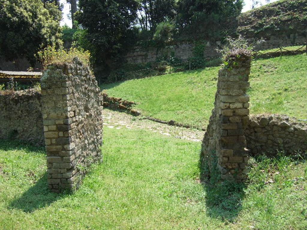 NGOF Pompeii. May 2006. View from area north-west of tomb looking towards the city walls near the Nola Gate.
According to De Caro – 
“The group of people found in the recent explorations was composed of 15 individuals, all of whom fell in the space of a few metres”.
See De Caro S., 1976. Scavi nell’area fuori Porta Nola a Pompei: Cronache Pompeiane V, (p.95-101).
The area where they were found can be seen, where the grassed area meets the roadway, looking through the gateway through which ran a beaten earth roadway.
The bodies were found in front of the gateway and down to and on the tufa block road. 
A beaten earth road ran through the gateway and joined with the tufa block road.
